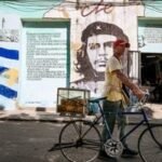 A man sells pastries in Havana, Cuba, in front of a mural of Che Guevara, symbolizing the enduring Cuban Revolution amidst new political pressures.