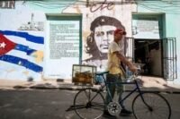 A man sells pastries in Havana, Cuba, in front of a mural of Che Guevara, symbolizing the enduring Cuban Revolution amidst new political pressures.