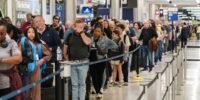 TSA agents working at an airport security checkpoint with travelers in the background.
