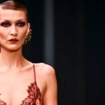 عارضة الأزياء بيلا حديد تتألق بإطلالة جريئة من الدانتيل الأحمر والأسود على منصة عرض سان لوران في أسبوع الموضة بباريس، مع مكياج غوثي وتسريحة شعر مشدودة.