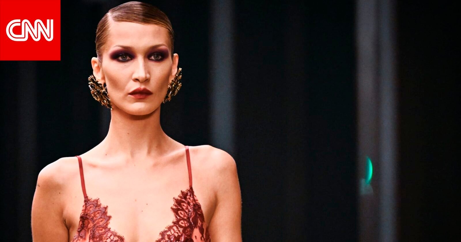 عارضة الأزياء بيلا حديد تتألق بإطلالة جريئة من الدانتيل الأحمر والأسود على منصة عرض سان لوران في أسبوع الموضة بباريس، مع مكياج غوثي وتسريحة شعر مشدودة.