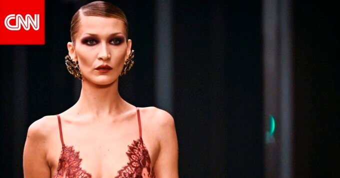 عارضة الأزياء بيلا حديد تتألق بإطلالة جريئة من الدانتيل الأحمر والأسود على منصة عرض سان لوران في أسبوع الموضة بباريس، مع مكياج غوثي وتسريحة شعر مشدودة.