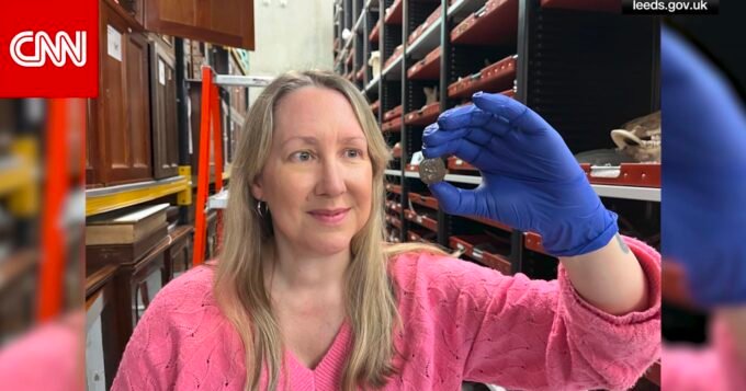 صورة مقربة لعملة معدنية قديمة ذات نقوش باهتة، مع خلفية لمتحف.