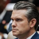 Secretary of Defense Pete Hegseth speaking at a press conference, with a blurred image of the US-Mexico border in the background.