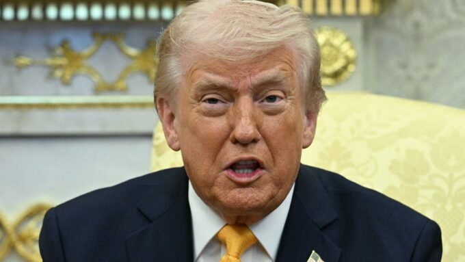 Former President Donald Trump speaking, with Federal Reserve Chair Jerome Powell in the background, symbolizing the ongoing conflict.