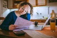 A person looking stressed while reviewing financial documents, with a calculator and bills on a table.