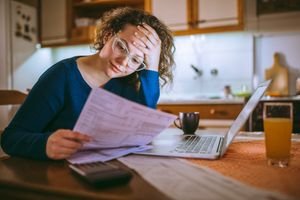 A person looking stressed while reviewing financial documents, with a calculator and bills on a table.