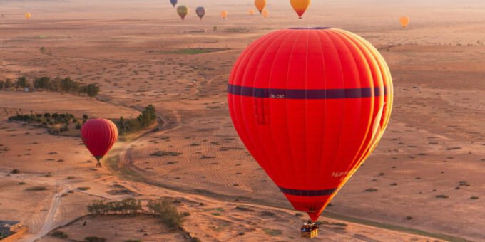 Des montgolfières colorées s'envolant au-dessus des paysages ocres de Marrakech, avec les montagnes de l'Atlas en arrière-plan, symbolisant le boom touristique et le besoin de régulation.