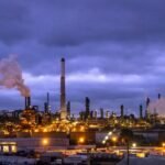 An aerial view of a refinery complex with pipelines, set against a backdrop of mountains and the ocean, symbolizing California's isolated energy infrastructure.