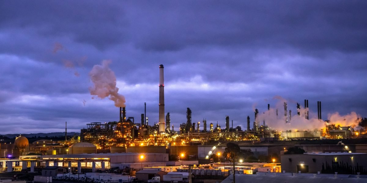An aerial view of a refinery complex with pipelines, set against a backdrop of mountains and the ocean, symbolizing California's isolated energy infrastructure.