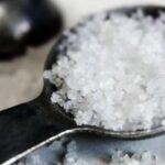 Une main saupoudrant du sel sur un plat, avec des symboles de danger et une balance pour illustrer l'équilibre nécessaire et les risques de l'excès.