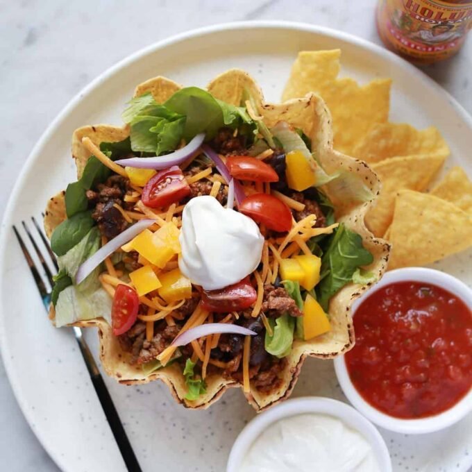 A vibrant homemade taco salad served in a crispy tortilla bowl, topped with ground meat, fresh vegetables, cheese, salsa, and sour cream.