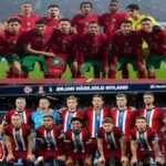 Joueurs de l'équipe nationale de football du Maroc célébrant un but lors d'un match international, avec le drapeau marocain en arrière-plan.