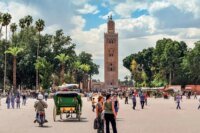 Vue aérienne d'une ville marocaine historique, avec des toits ocres et des palmiers, sous un ciel bleu.