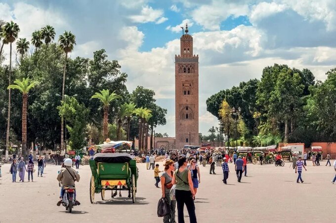 Vue aérienne d'une ville marocaine historique, avec des toits ocres et des palmiers, sous un ciel bleu.