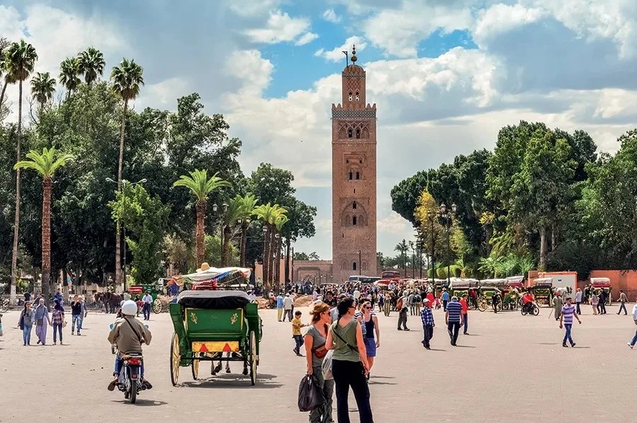 Vue aérienne d'une ville marocaine historique, avec des toits ocres et des palmiers, sous un ciel bleu.