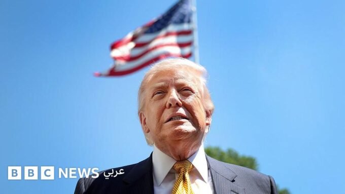 صورة للرئيس الأمريكي دونالد ترامب يتحدث في مؤتمر صحفي، مع خلفية تظهر أعلام الولايات المتحدة.