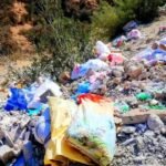 صورة لغابة أغروض مليئة بالنفايات البلاستيكية ومخلفات البناء، مع أشجار أركان متضررة في الخلفية.