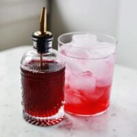 A glass bottle of vibrant red homemade cherry syrup next to a bowl of fresh cherries and a cocktail glass.