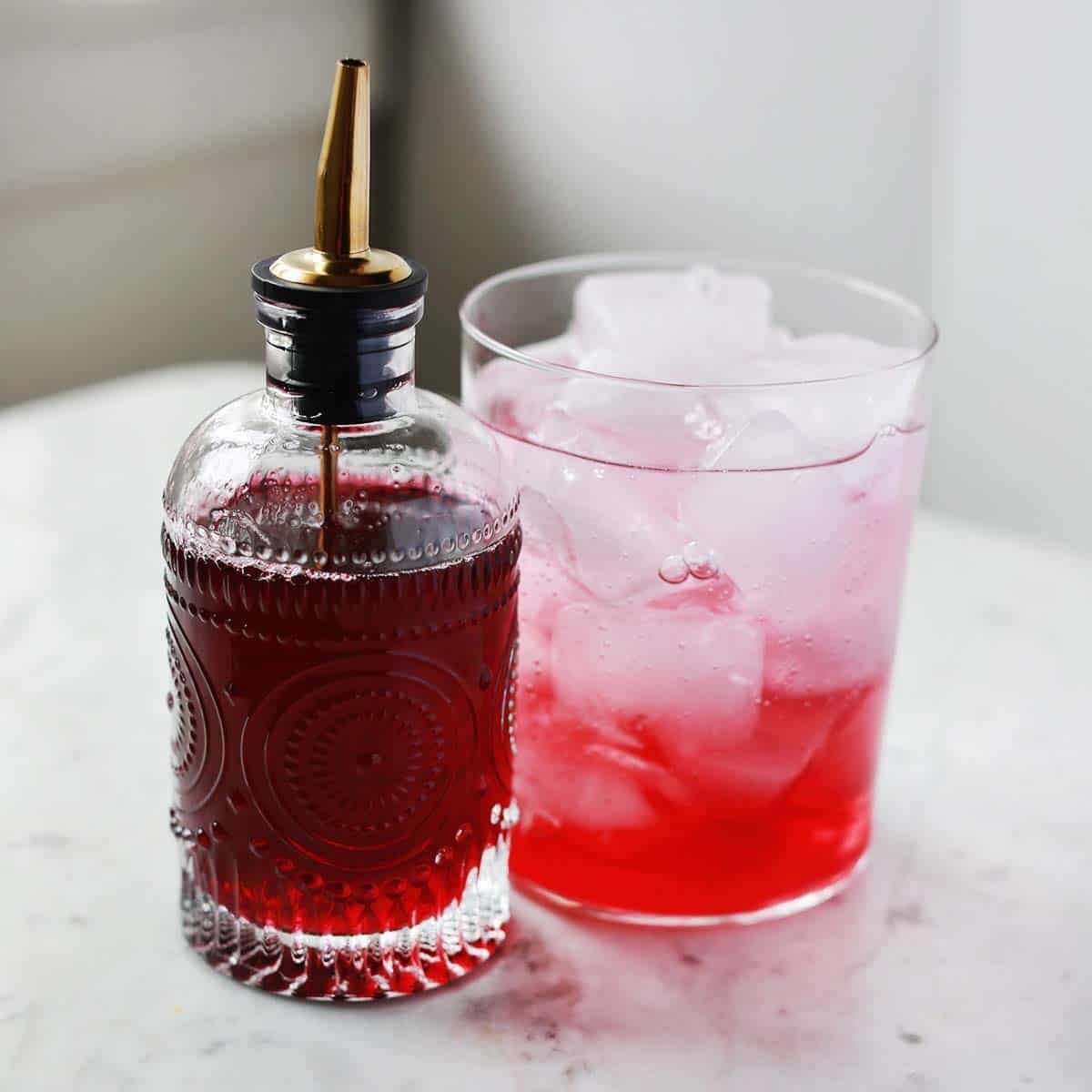 A glass bottle of vibrant red homemade cherry syrup next to a bowl of fresh cherries and a cocktail glass.
