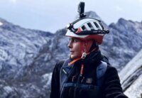 Nawal Sfendla, alpiniste marocaine, souriante et équipée pour l'alpinisme, avec un paysage montagneux en arrière-plan, symbolisant sa détermination et son exploit sur l'Everest et le Lhotse.