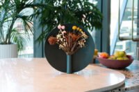 The Nimbus Vase by Ben Peng, a sculptural vessel with a metallic halo-like ring framing flowers, set in a minimalist interior.