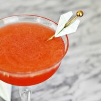 A vibrant orange Paper Plane cocktail served in a coupe glass, garnished with a tiny paper airplane.