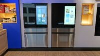 A modern Samsung Bespoke refrigerator with a large touchscreen display showing various smart features and food recognition.