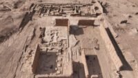 Vue aérienne des fouilles archéologiques de Sijilmassa, montrant les vestiges de la cité médiévale et la structure métallique moderne en construction, évoquant les dunes du désert.