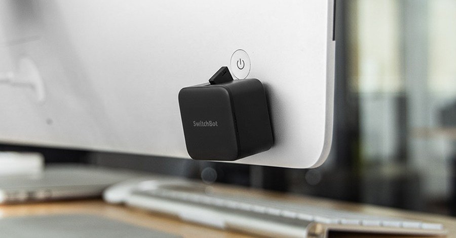 A SwitchBot Bot Rechargeable device, a small white robotic arm, attached to a black monitor button, ready to press it.