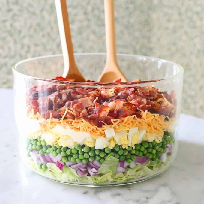 A beautifully presented 7 Layer Salad in a clear glass bowl, showcasing distinct layers of lettuce, peas, eggs, cheese, and bacon.