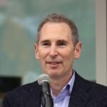 Amazon CEO Andy Jassy speaking, with a subtle AI-themed background.