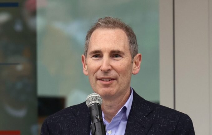 Amazon CEO Andy Jassy speaking, with a subtle AI-themed background.