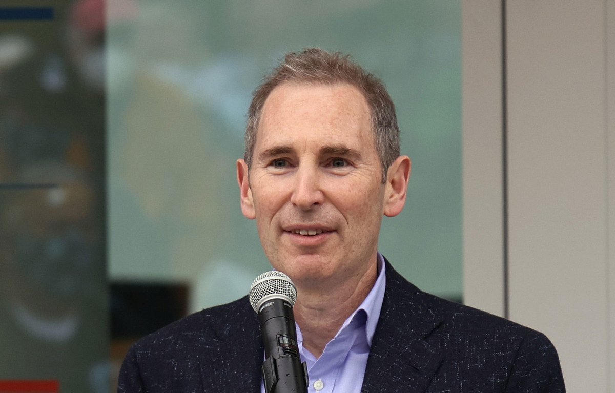 Amazon CEO Andy Jassy speaking, with a subtle AI-themed background.