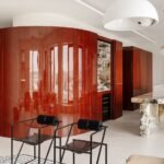 A high-gloss oval volume in deep red Merbau veneer at the center of a modern, open-plan apartment, reflecting natural light.