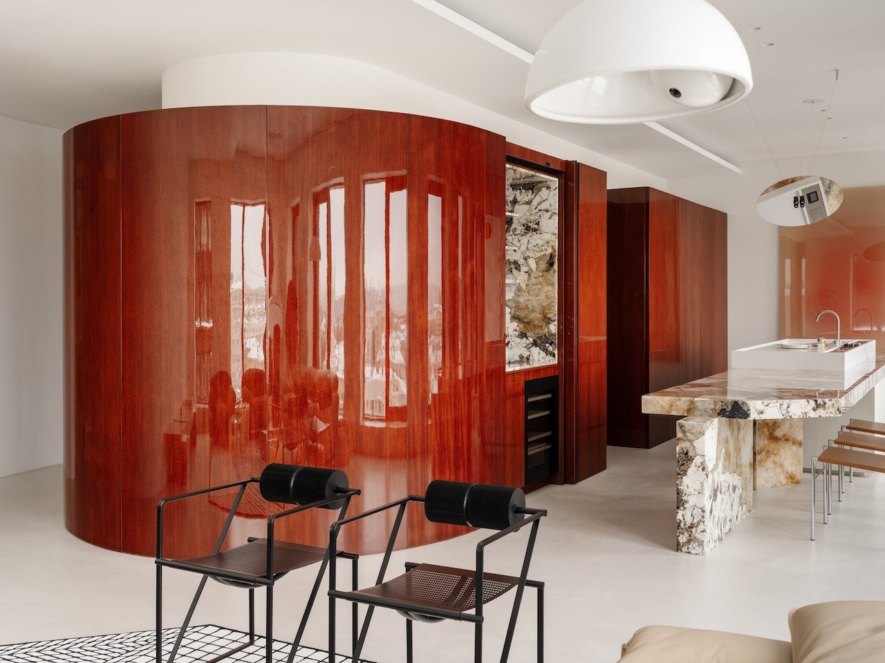 A high-gloss oval volume in deep red Merbau veneer at the center of a modern, open-plan apartment, reflecting natural light.