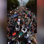 صورة لمشجعين مكسيكيين يؤدون الموجة البشرية في ملعب مزدحم أو شارع رئيسي، يظهرون حماسًا كبيرًا.
