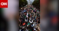 صورة لمشجعين مكسيكيين يؤدون الموجة البشرية في ملعب مزدحم أو شارع رئيسي، يظهرون حماسًا كبيرًا.
