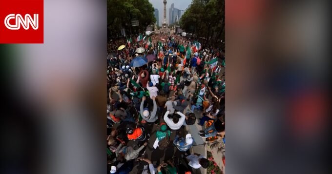 صورة لمشجعين مكسيكيين يؤدون الموجة البشرية في ملعب مزدحم أو شارع رئيسي، يظهرون حماسًا كبيرًا.