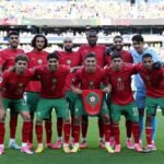 Joueurs de l'équipe nationale marocaine célébrant un but lors de la Coupe du Monde 2026, avec le drapeau du Maroc en arrière-plan.