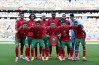 Joueurs de l'équipe nationale marocaine célébrant un but lors de la Coupe du Monde 2026, avec le drapeau du Maroc en arrière-plan.
