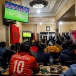 Des supporters de football regardant un match de la Coupe du Monde 2026 dans un café marocain, avec des écrans de télévision affichant un match de football et des personnes buvant du thé ou du café.