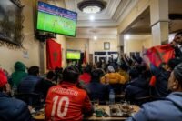 Des supporters de football regardant un match de la Coupe du Monde 2026 dans un café marocain, avec des écrans de télévision affichant un match de football et des personnes buvant du thé ou du café.