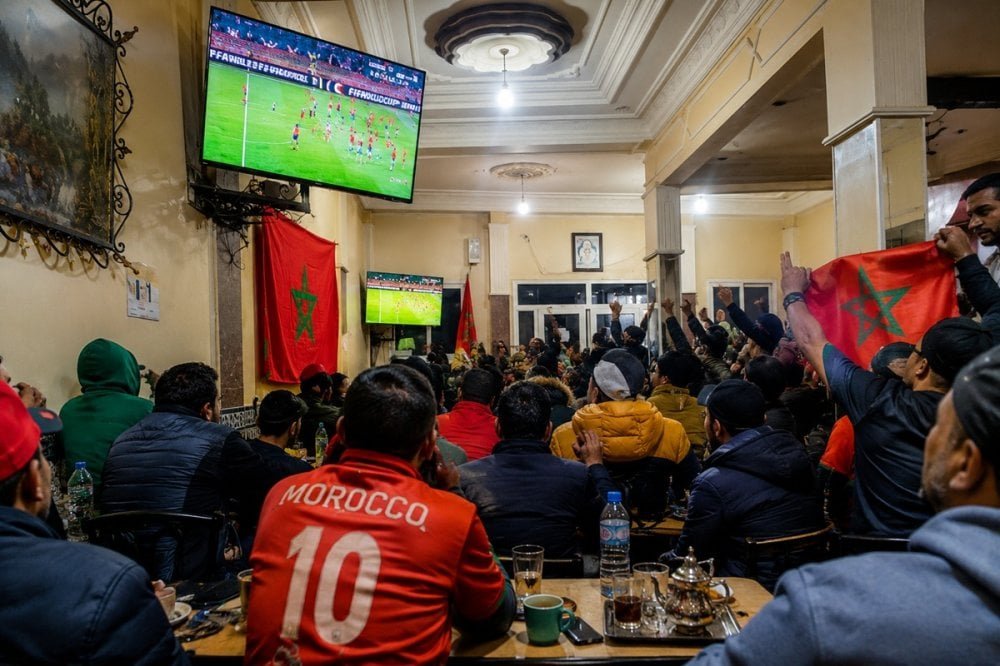 Des supporters de football regardant un match de la Coupe du Monde 2026 dans un café marocain, avec des écrans de télévision affichant un match de football et des personnes buvant du thé ou du café.