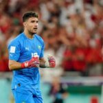 Portrait de Munir El Kajoui, gardien de but de l'équipe nationale marocaine, les Lions de l'Atlas, en tenue de match, regardant vers l'avant avec détermination. Le fond est flou, mettant en valeur le joueur.