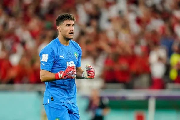 Portrait de Munir El Kajoui, gardien de but de l'équipe nationale marocaine, les Lions de l'Atlas, en tenue de match, regardant vers l'avant avec détermination. Le fond est flou, mettant en valeur le joueur.