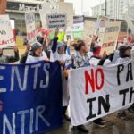 Protesters in hospital gowns holding 'Hands Off Our NHS' signs outside a conference, demonstrating against Palantir's data deal.
