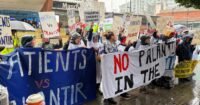 Protesters in hospital gowns holding 'Hands Off Our NHS' signs outside a conference, demonstrating against Palantir's data deal.