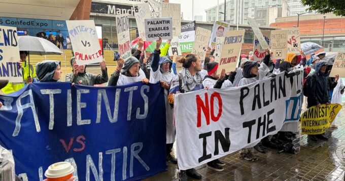 Protesters in hospital gowns holding 'Hands Off Our NHS' signs outside a conference, demonstrating against Palantir's data deal.