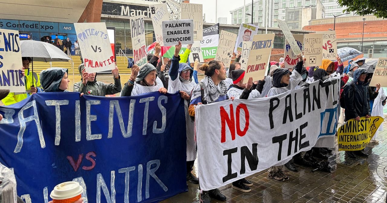 Protesters in hospital gowns holding 'Hands Off Our NHS' signs outside a conference, demonstrating against Palantir's data deal.